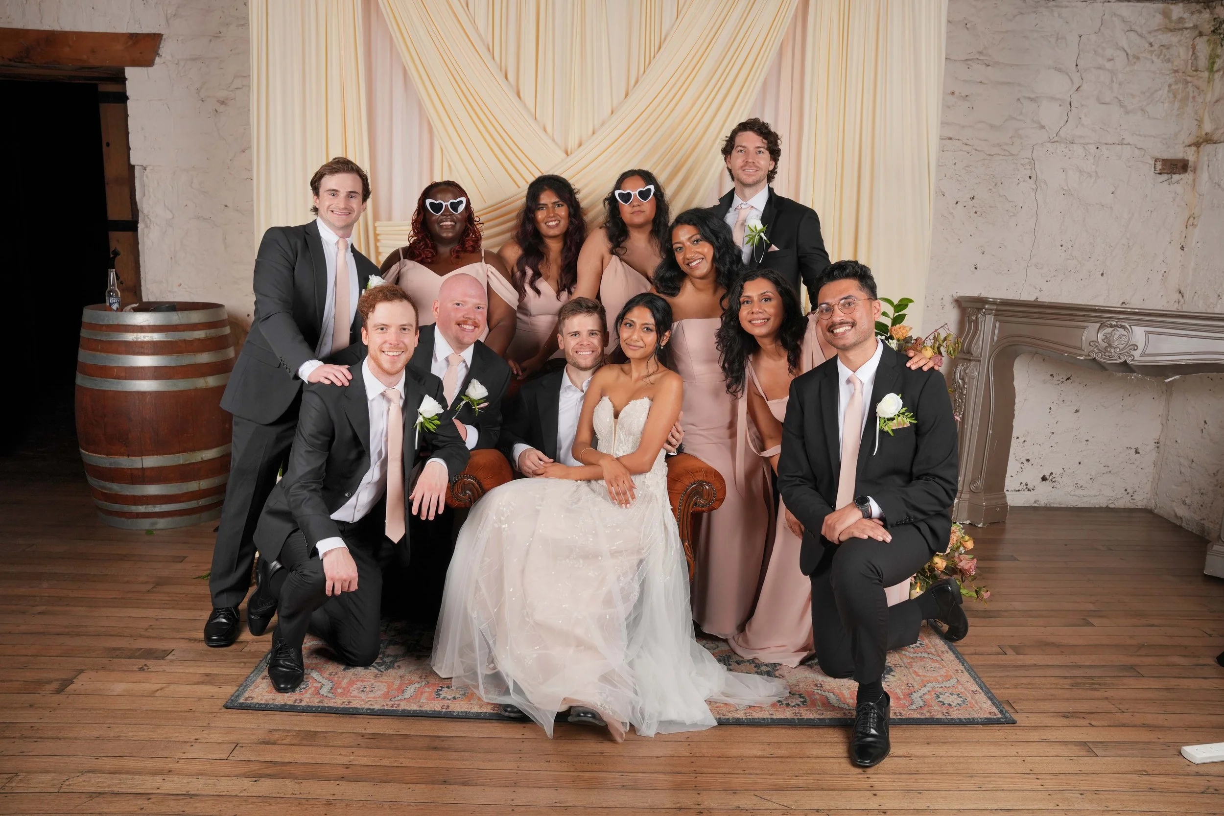 Group of people at a wedding celebration, with the bride in a white gown seated in the center, surrounded by bridesmaids in pink dresses, groomsmen in black suits, and other guests posing together in front of a draped backdrop.