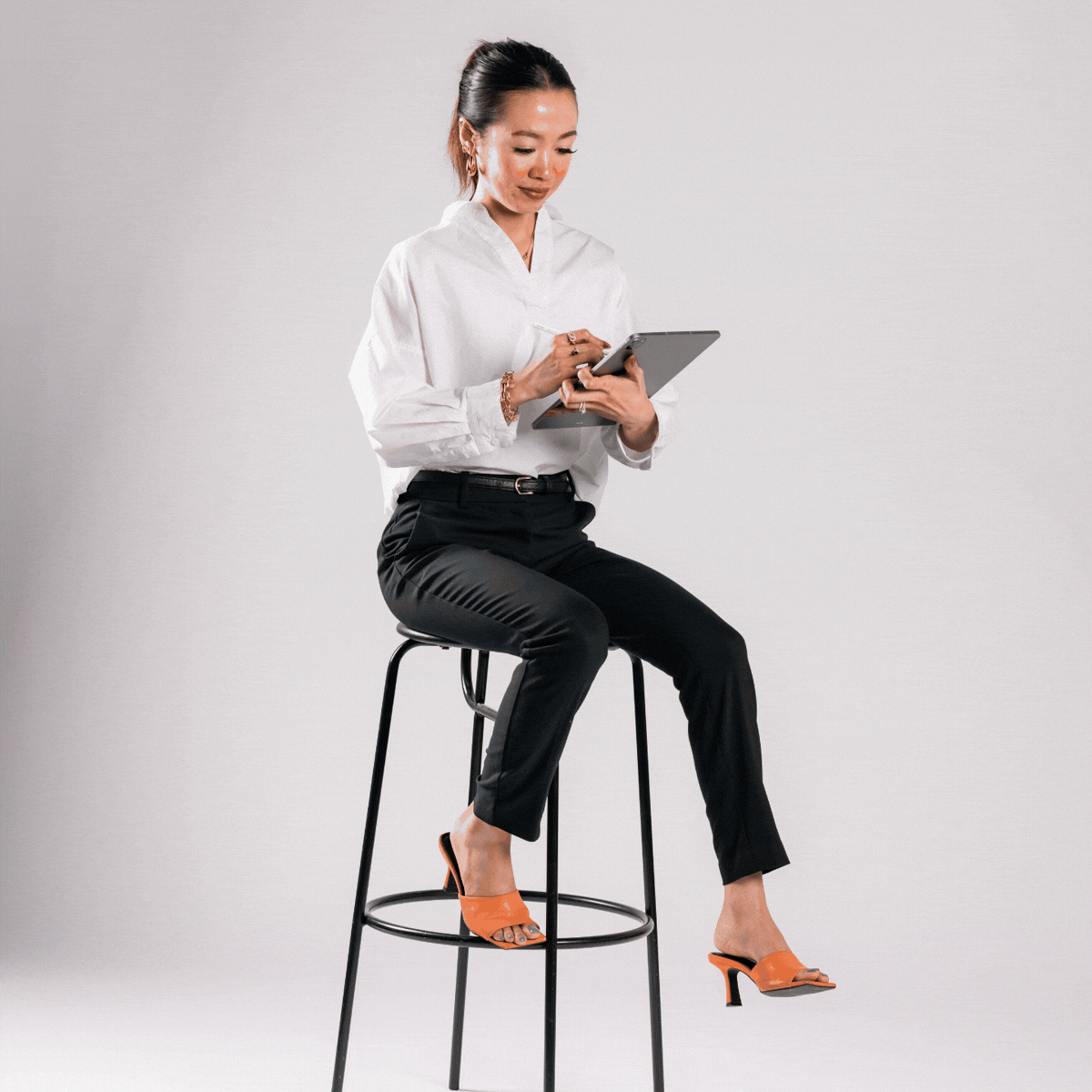 A woman sitting on a stool, dressed in business attire, using a tablet against a plain gray background.