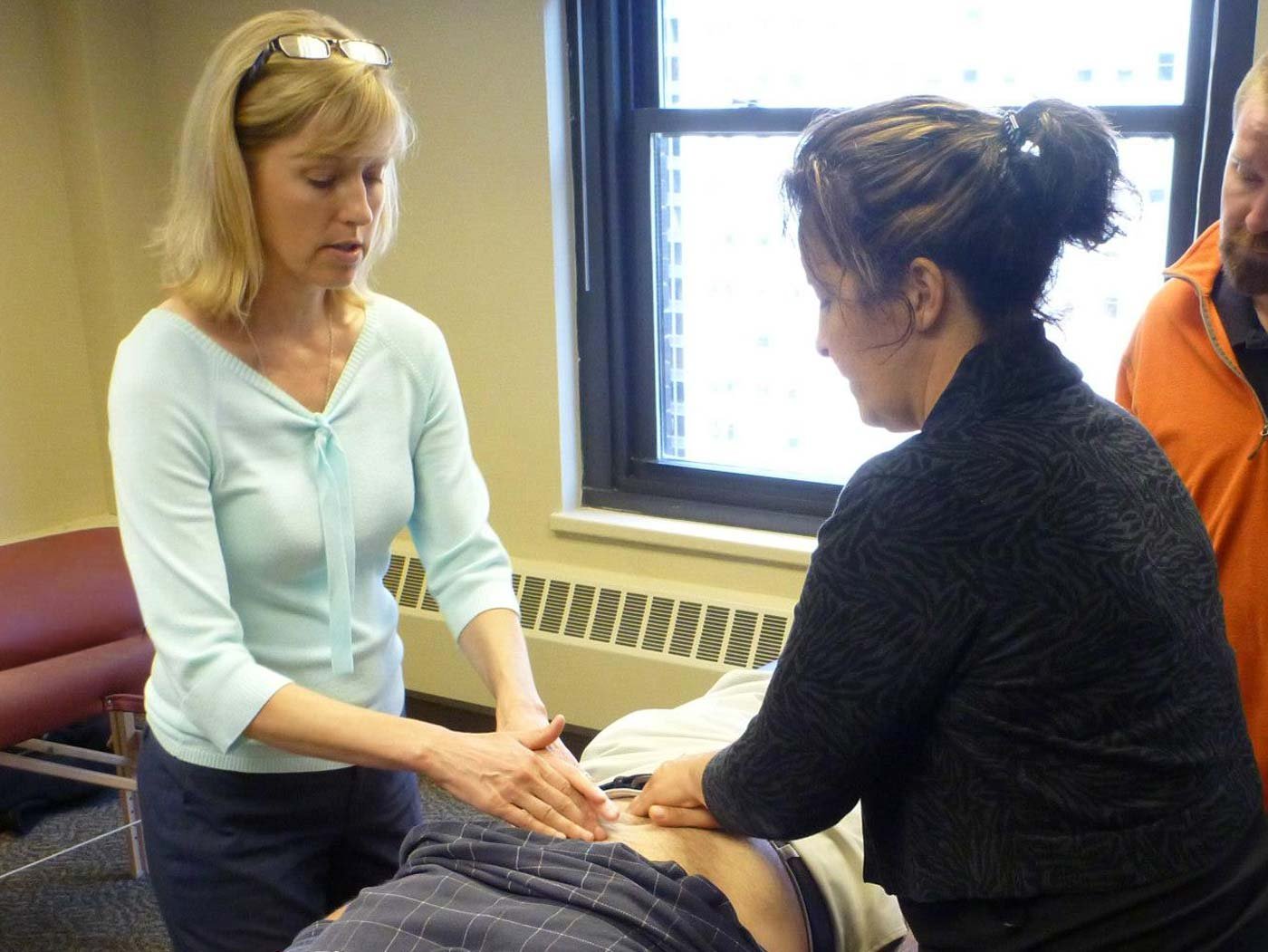 Tracy Thorne teaches students in a medical training or emergency response scenario. One woman is palpating a person lying on a table, while another woman and a man observe. The setting appears to be a training room with a window in the background.