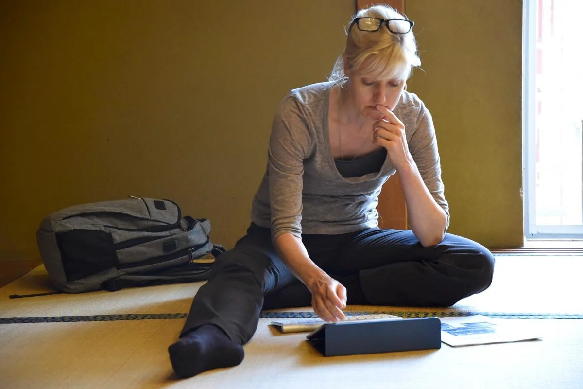 Tracy Thorne is sitting on the floor near a window, wearing glasses on her head, studying the information on a tablet with her hand near her mouth. There is a backpack beside her.