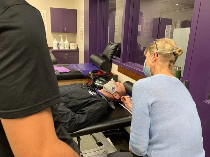 Tracy Thorne is wearing a blue cardigan and face mask while she interacts with a patient lying on a medical examination table in a clinical setting with purple walls. She demonstrates moxibustion and acupuncture techniques.