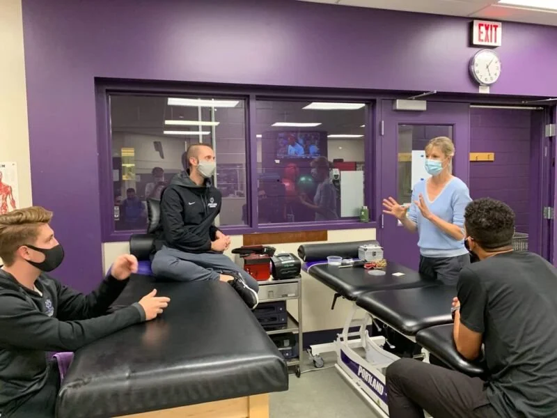 Four sports physical therapists in a medical training room, all wearing masks. Tracy is standing near a medical table, teaching acupuncture and moxibustion techniques to three seated students, with a purple wall and large window behind her.