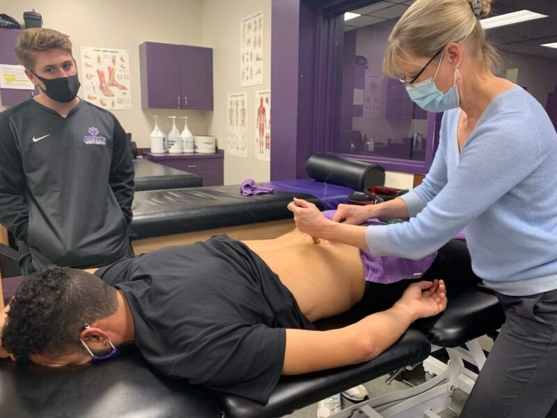 Tracy Thorne conducts an examination on a patient lying face down on a medical table, with another person observing in the background in a clinical setting.