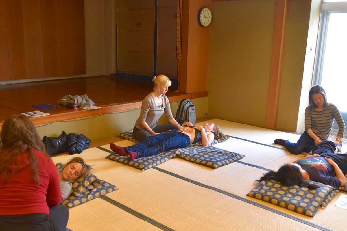 A group of people practicing abdominal palpation techniques on their partners lying on cushions on tatami mats in a room with wooden walls and a large window.