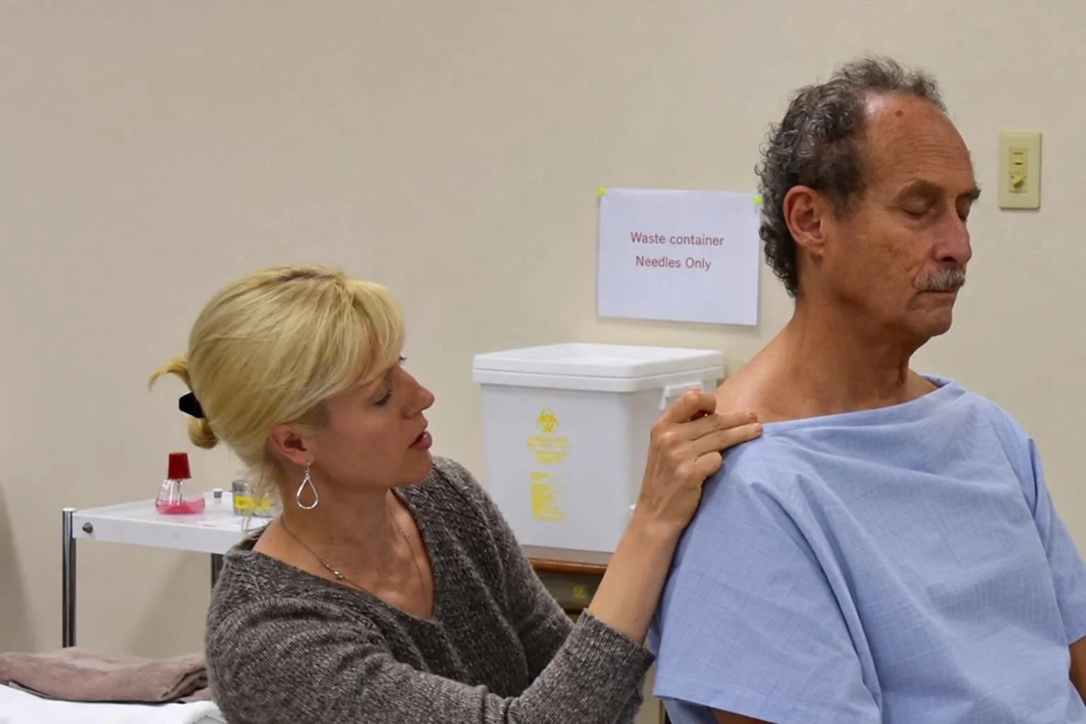 A woman with blonde hair and earrings is administering an injection to an older man with gray hair, in a medical setting. The man is seated, with his eyes closed, wearing a blue gown. There is a white container labeled "Waste container Needles Only" 