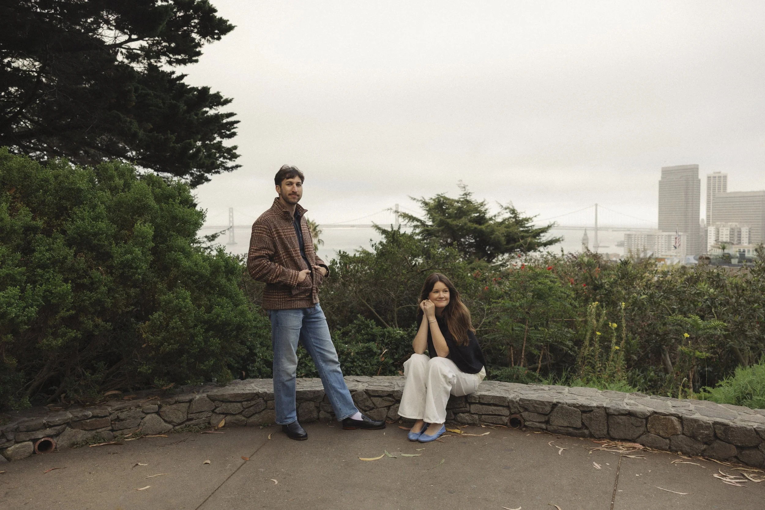 A man with dark hair and beard standing with his hands in his pockets, and a woman with long red hair sitting on a low stone wall, both surrounded by greenery with trees and bushes, and a cityscape with high-rise buildings and a bridge in the background on a cloudy day.