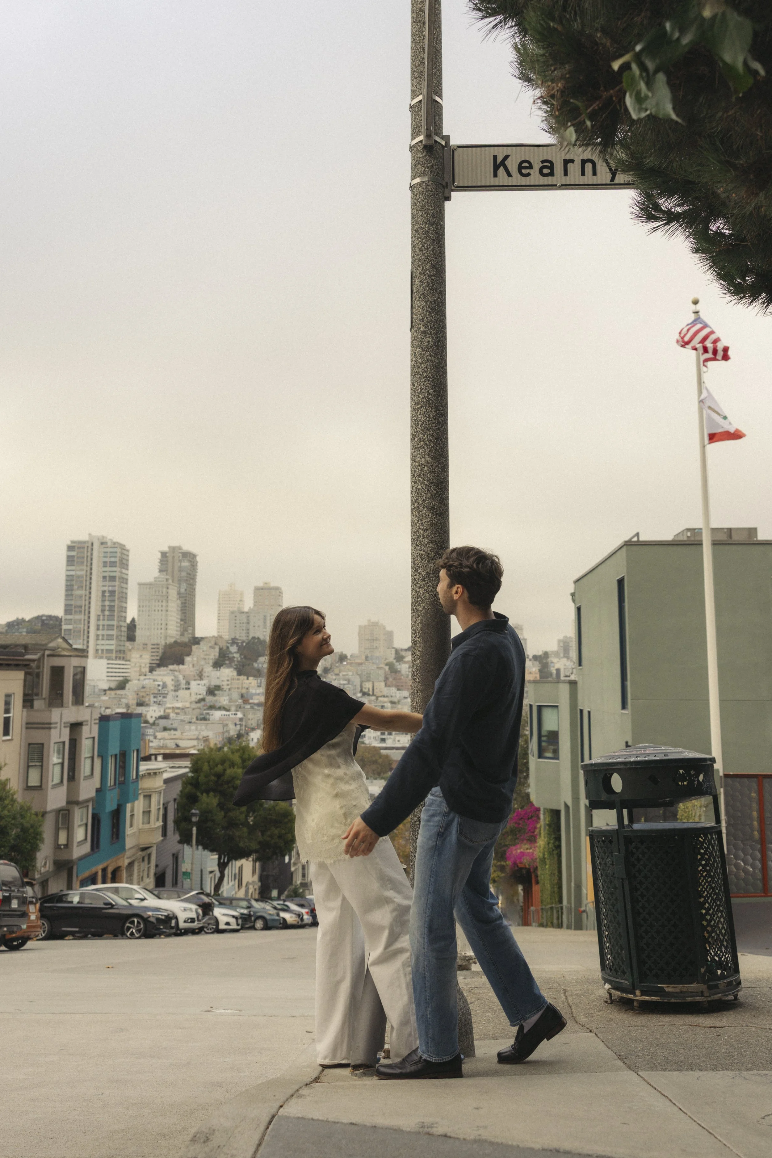 A couple holding hands and facing each other outdoors in front of a cityscape with tall buildings. The woman is wearing white pants and a black top, and the man is dressed in jeans and a dark jacket. They are standing on a sidewalk near a parking lot and a trash bin, with trees and American flags visible in the background.