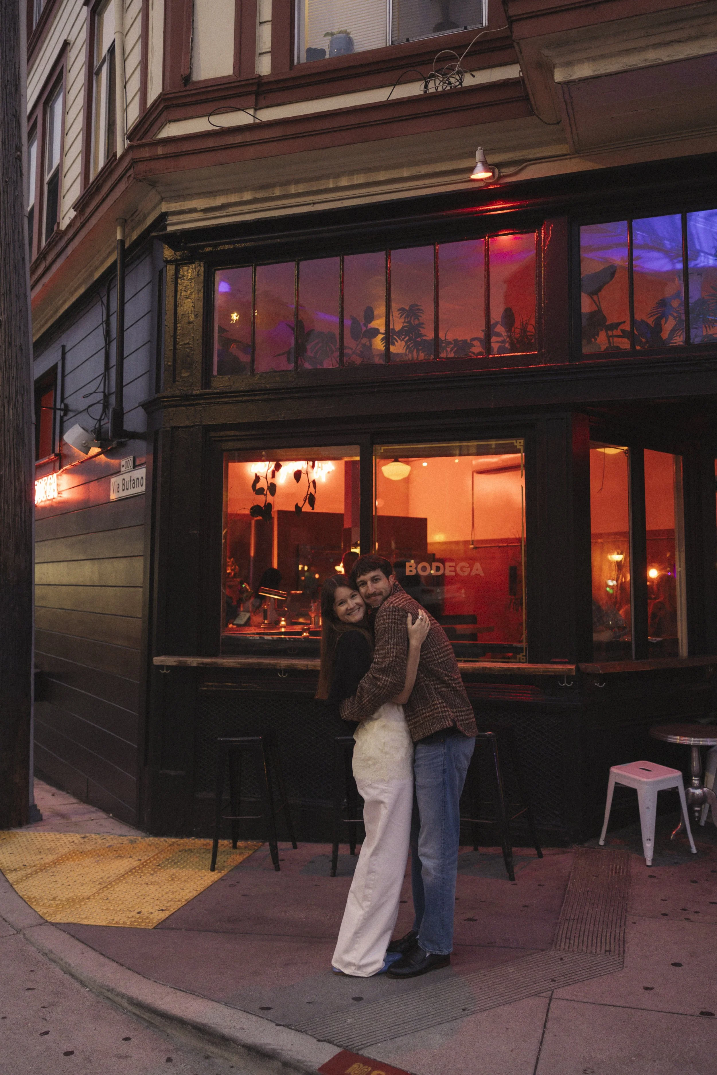 A couple hugging outside a restaurant at dusk, with a building featuring large windows and colorful lighting in the background.