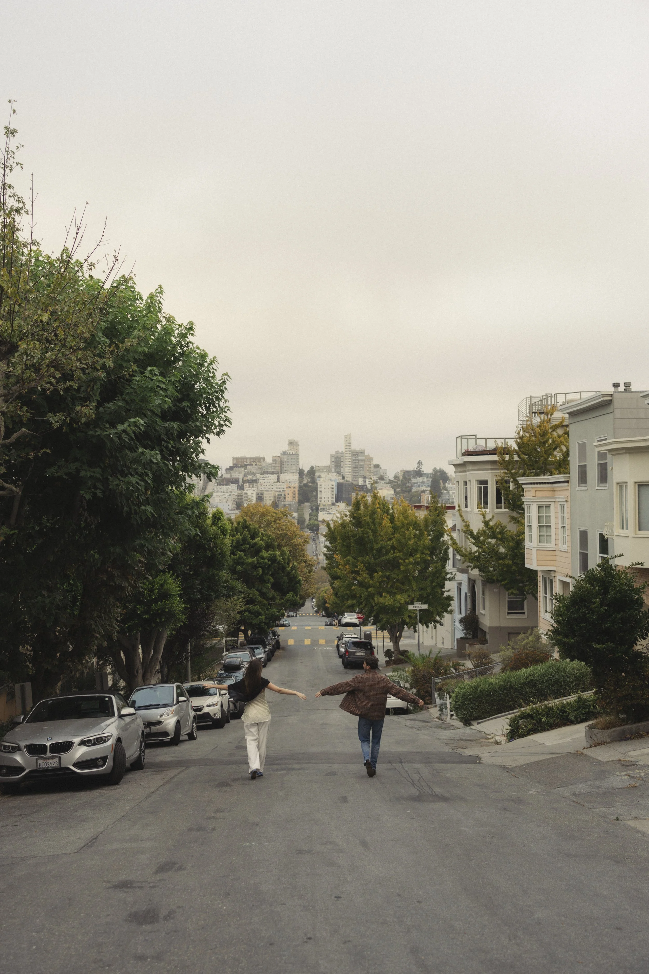 Two people walking down a city street on a slightly inclined hill, holding hands and stretching their arms out, with parked cars on either side and residential buildings in the background.