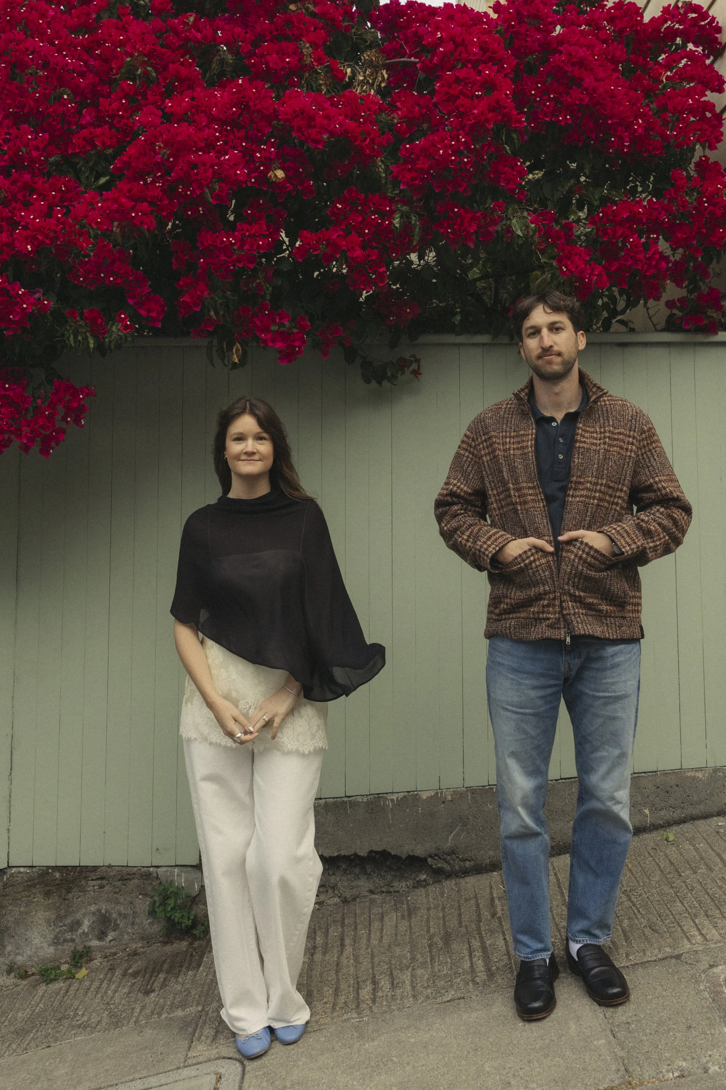 A woman and a man standing in front of a green fence with a large bush of red flowers above them. The woman is wearing a black top and cream-colored pants, and the man is wearing a brown patterned jacket with blue jeans.