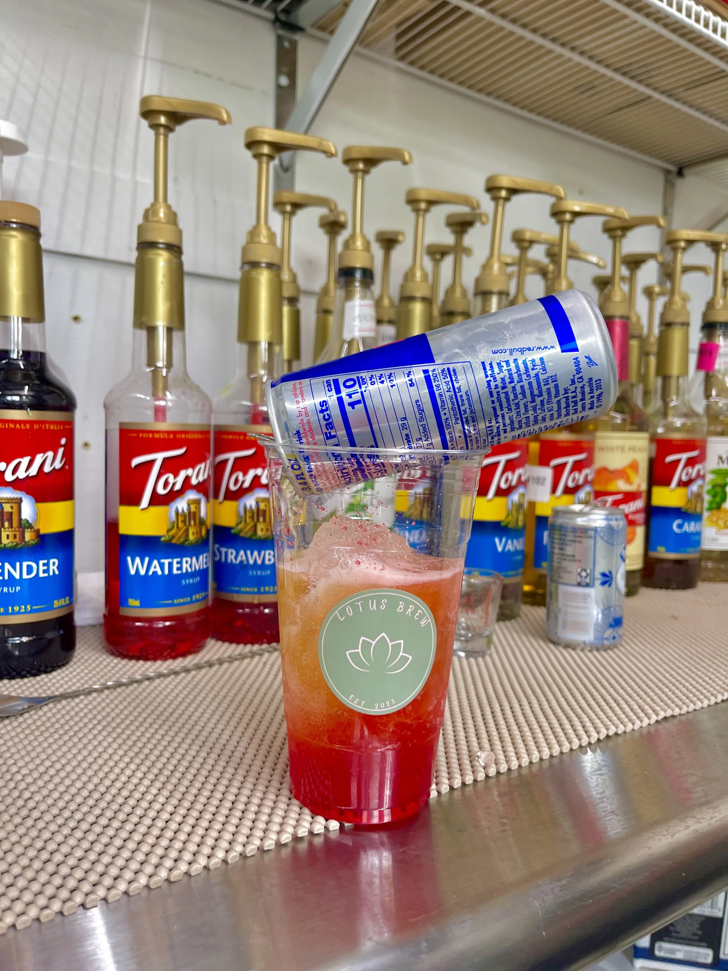 A plastic cup with a transparent straw containing a red and orange layered drink, placed on a metallic surface in front of a shelf with various syrups and dispensing pumps.