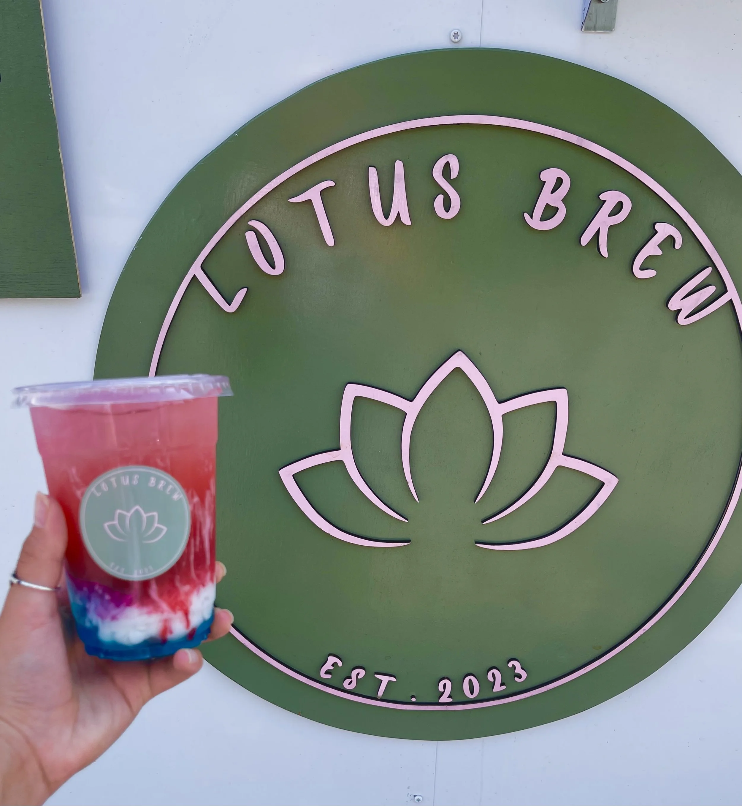 A person holding a pink and blue smoothie with whipped cream in front of a green circular sign that reads "Lotus Brooklyn" and "Est. 2023." The person's hand has rings.