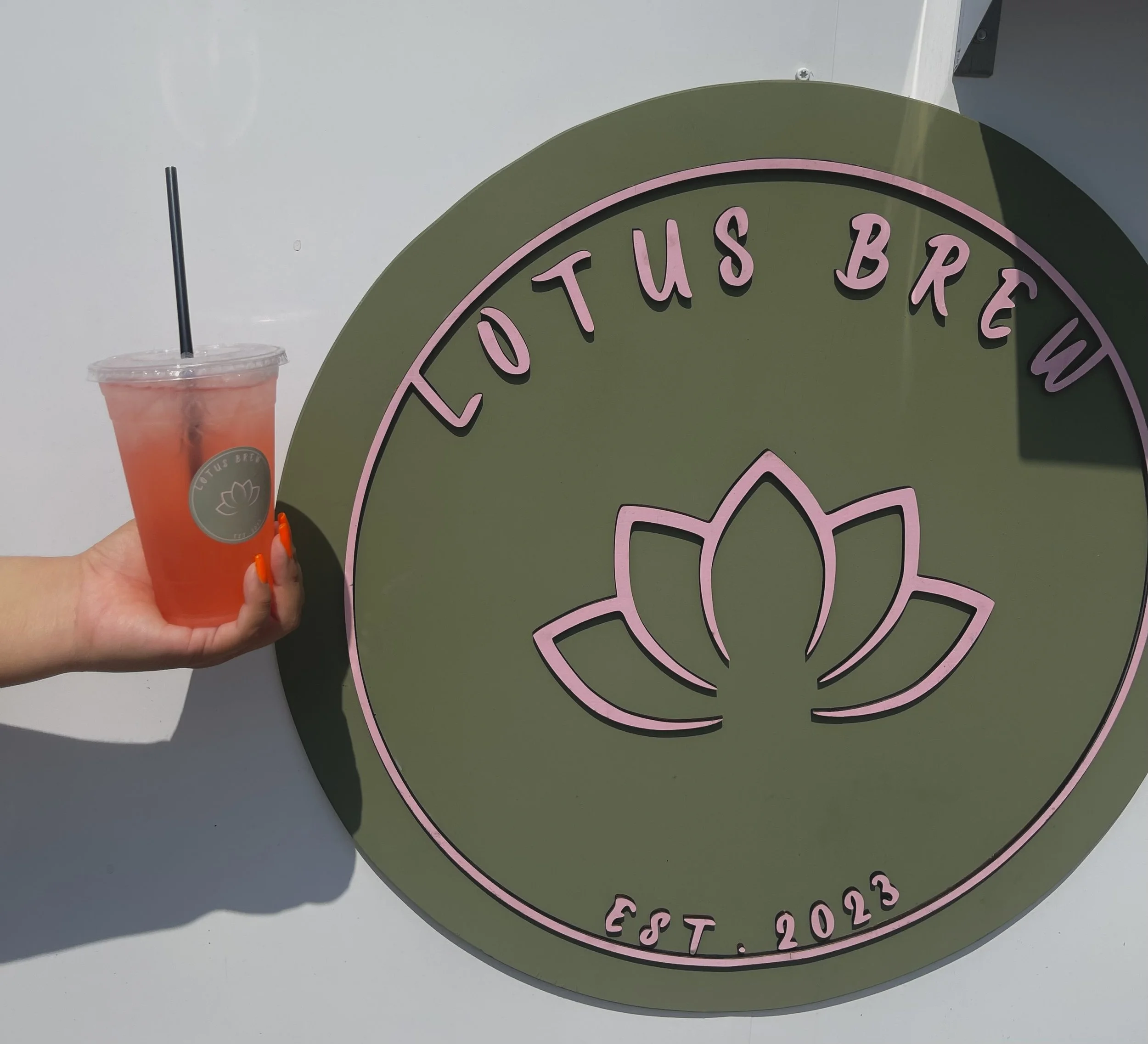 Person holding a pink drink in front of a green sign with pink lettering displaying "Lotus Brew" and the phrase "Est. 2023."