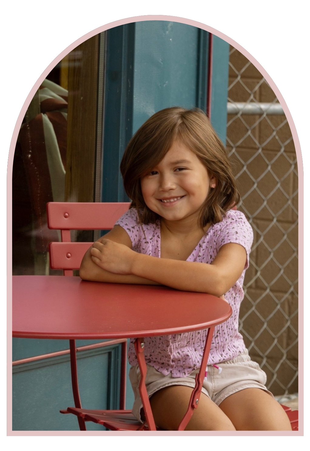 A young girl with short brown hair, smiling and sitting at a pink table at an outdoor cafe or patio, with a chain-link fence in the background.