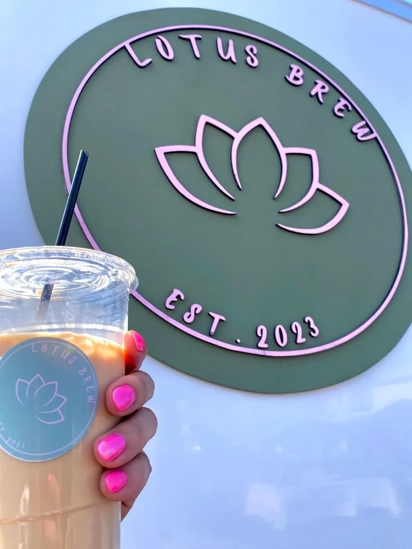 A hand holding a cup of iced beverage with pink nails in front of a green sign that reads "Lotus Brew Est. 2023" with a lotus flower logo.