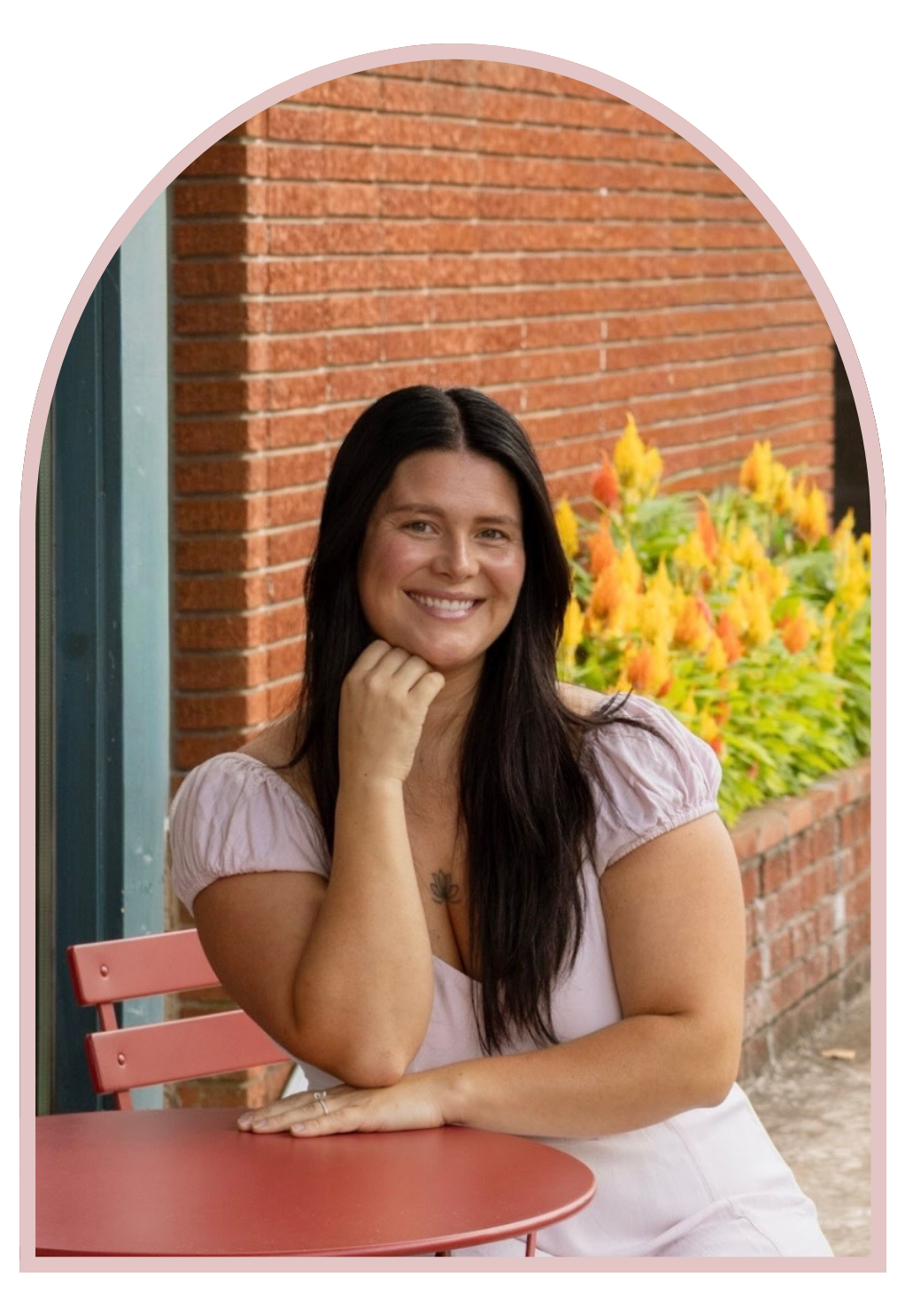 A woman with long dark hair smiling and resting her chin on her hand, sitting at a pink table outside near a red brick wall and yellow-orange flowers.