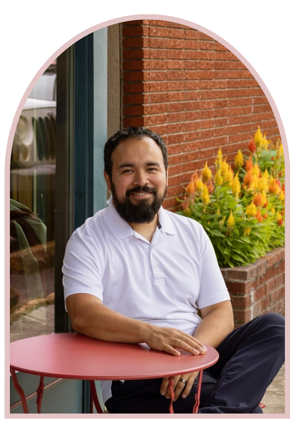 A man with a beard and dark hair sitting outside at a small round pink table, wearing a white polo shirt, with a brick wall and colorful flowers in the background.