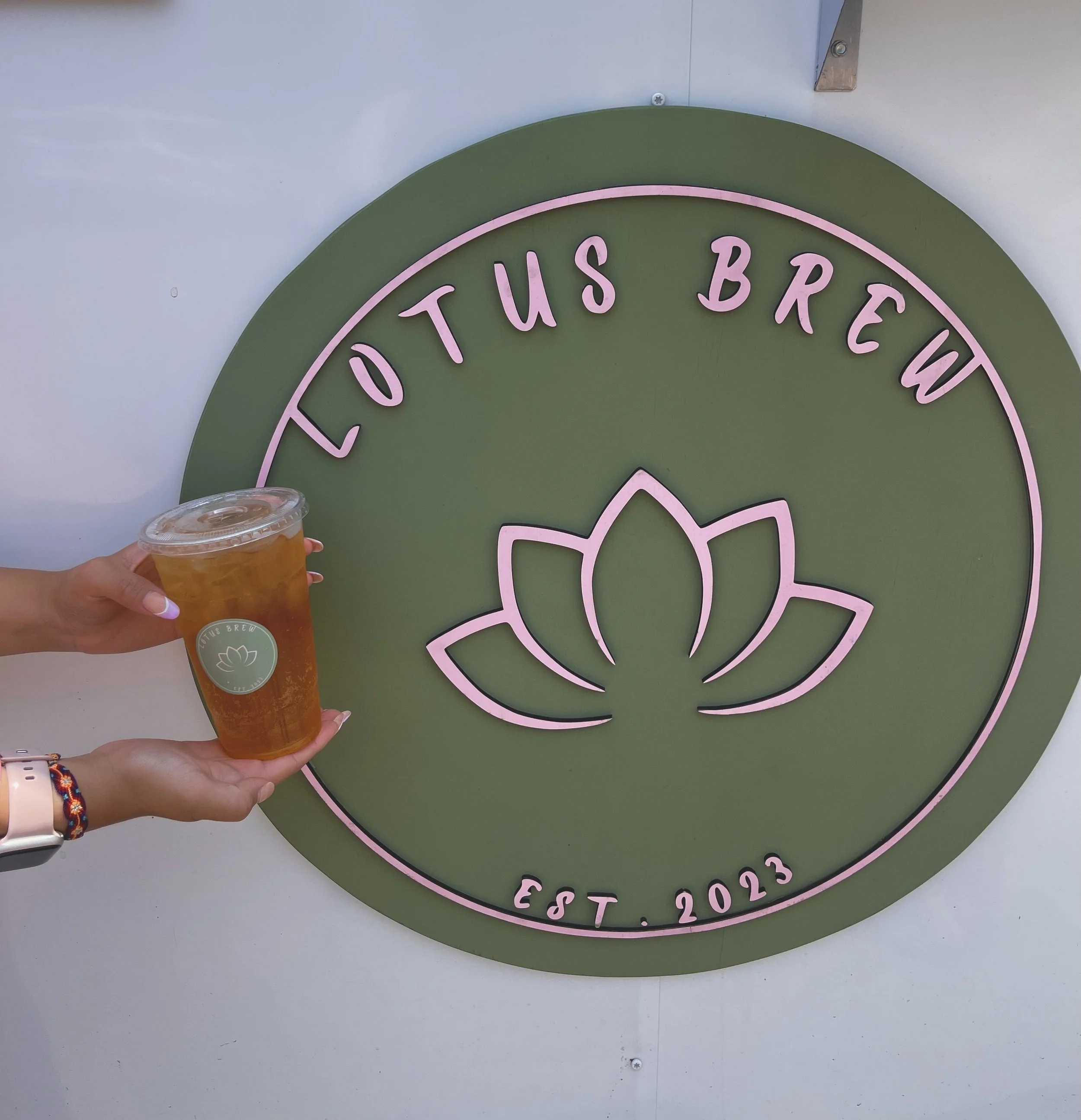 Hand holding a plastic cup of iced tea with a straw in front of a green and pink sign that reads "Lotus Brew" and "EST. 2023".
