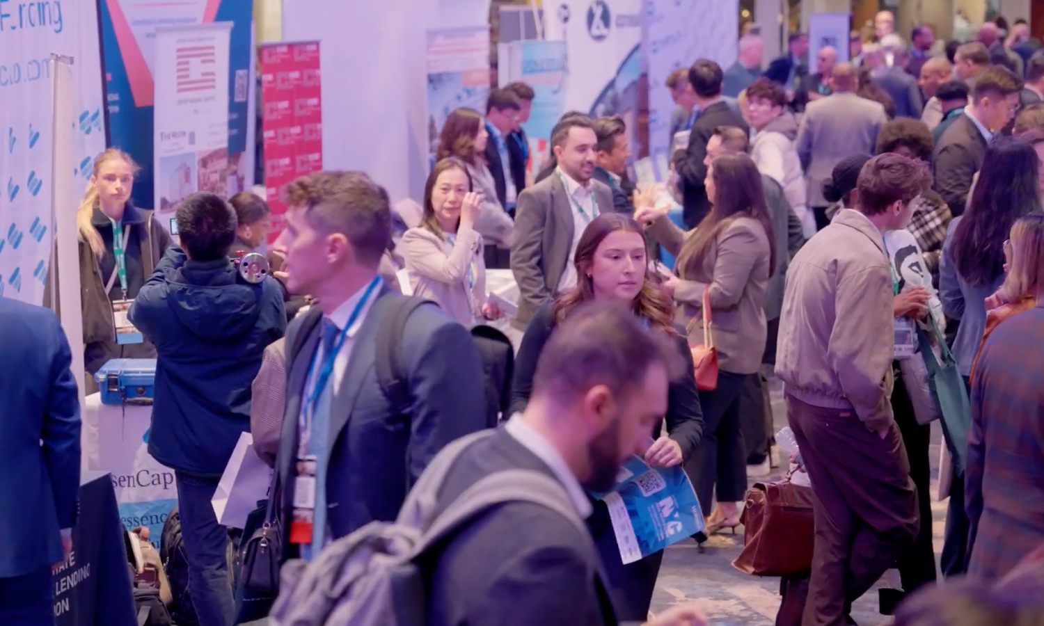 Crowd of people at a busy conference or trade show with various booths and displays in the background.