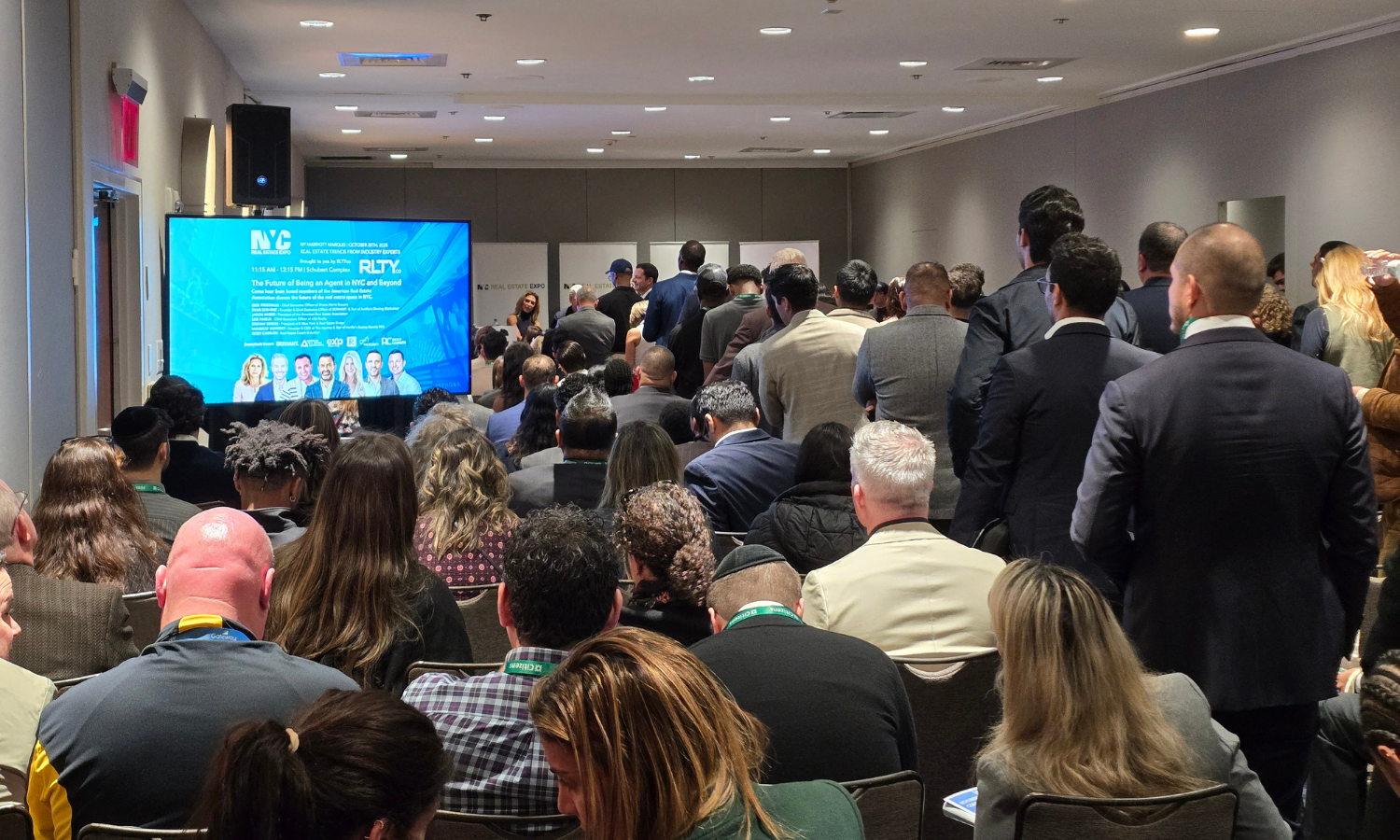 A large group of predominantly professionally dressed people attending a conference or seminar in a hotel conference room, with some standing and some seated, facing a stage with a large digital display screen showing an event related to real estate 