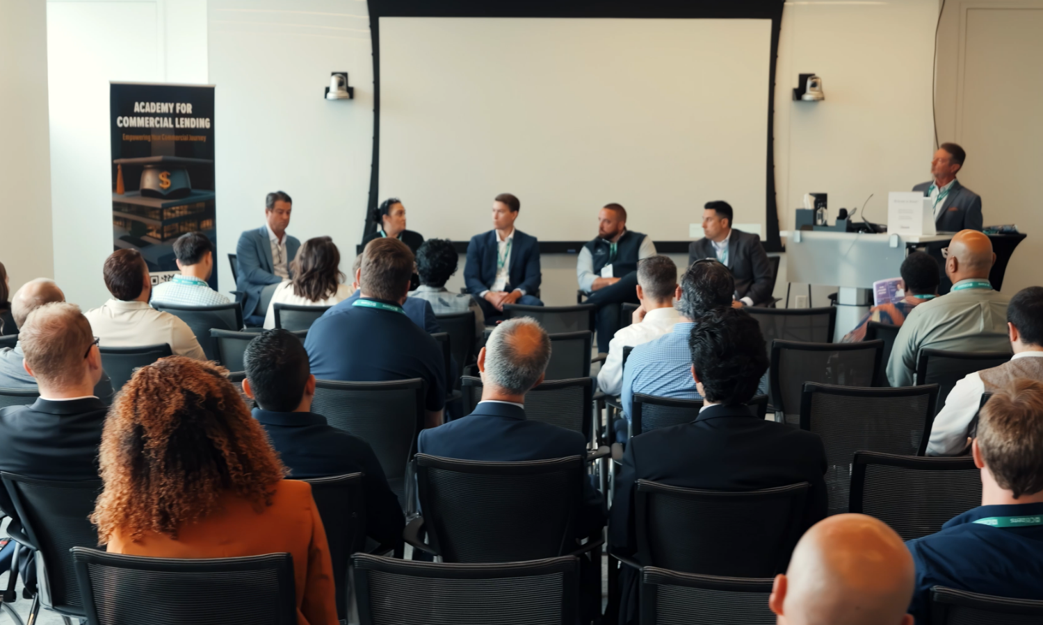 A conference or panel discussion with six speakers seated at the front and an audience of diverse individuals listening attentively. There is a large white screen behind the speakers and a banner on the left that reads 'Academy for Commercial Lending