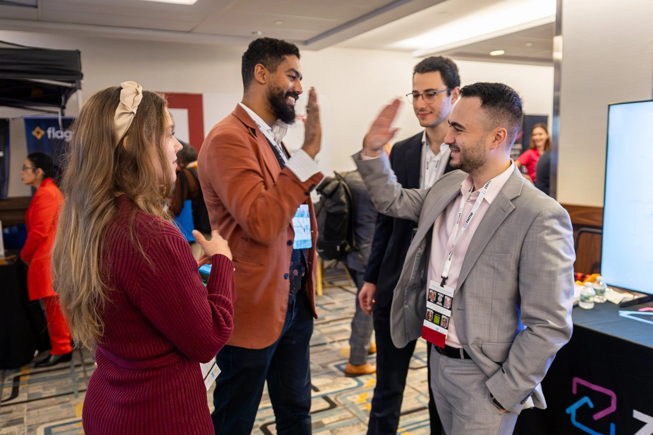 Four people are interacting in a conference or networking event. Two men and a woman are engaged in conversation, giving high-fives and smiling. The woman has long, wavy blonde hair and is wearing a red sweater. One man has a beard, is wearing glasses and a light gray suit. The other man has darker curly hair, a beard, and is wearing a reddish-brown jacket. They are all wearing conference badges.