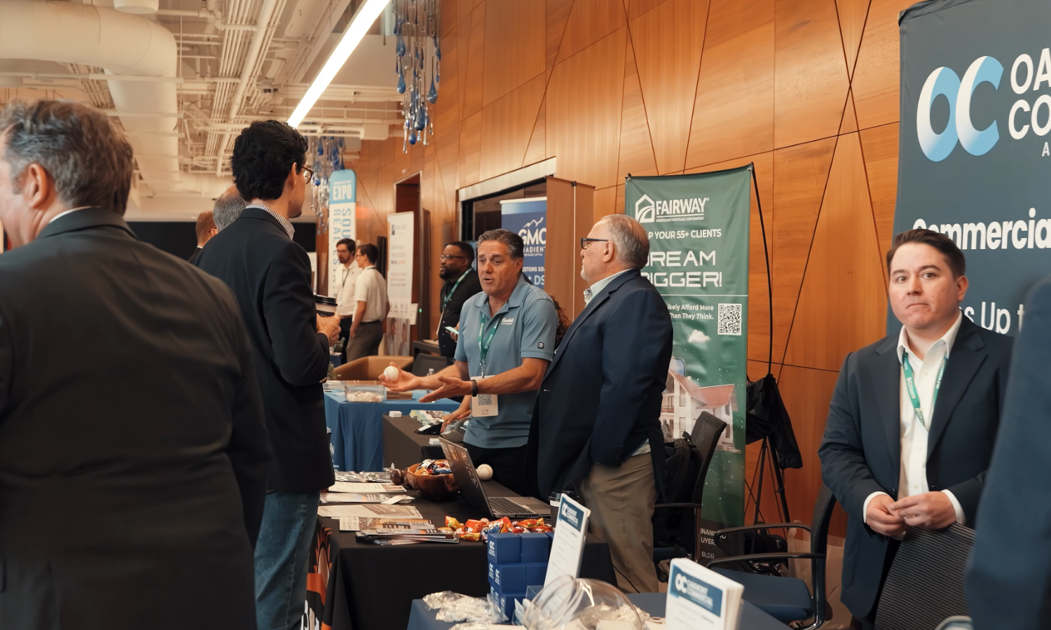 Business expo scene with multiple people at booths, including a man in a blue shirt talking to attendees, and banners advertising various companies.