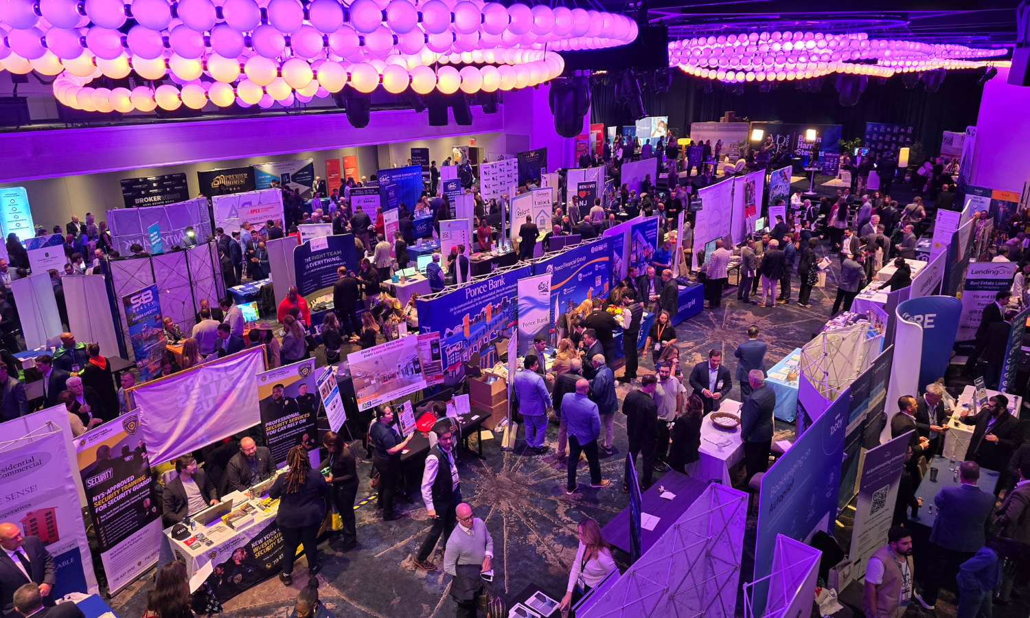 A busy conference or expo hall filled with numerous booths and attendees. The room is illuminated with purple lighting from large ceiling fixtures. People are walking, talking, and exploring various displays.