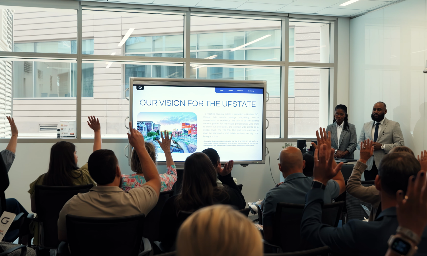 Business presentation in a conference room with attendees raising their hands, a digital screen displaying 'Our Vision for the Upstate', and two speakers standing at the front.