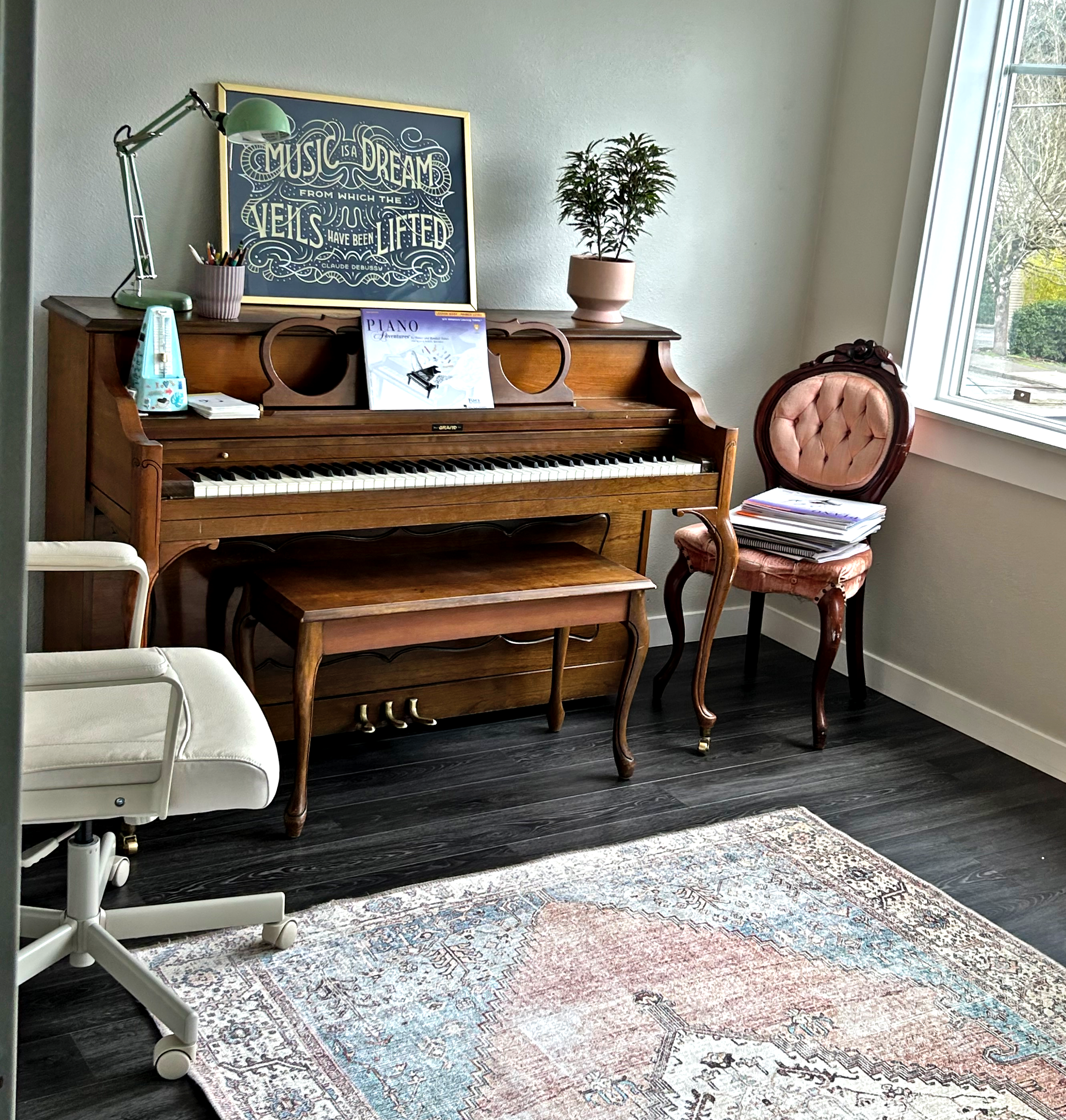 A room with a wooden upright piano, a framed quote art print, a potted plant, a chair with pink upholstery, a white medical chair, and a rug on a dark wood floor. Large window shows trees outside.