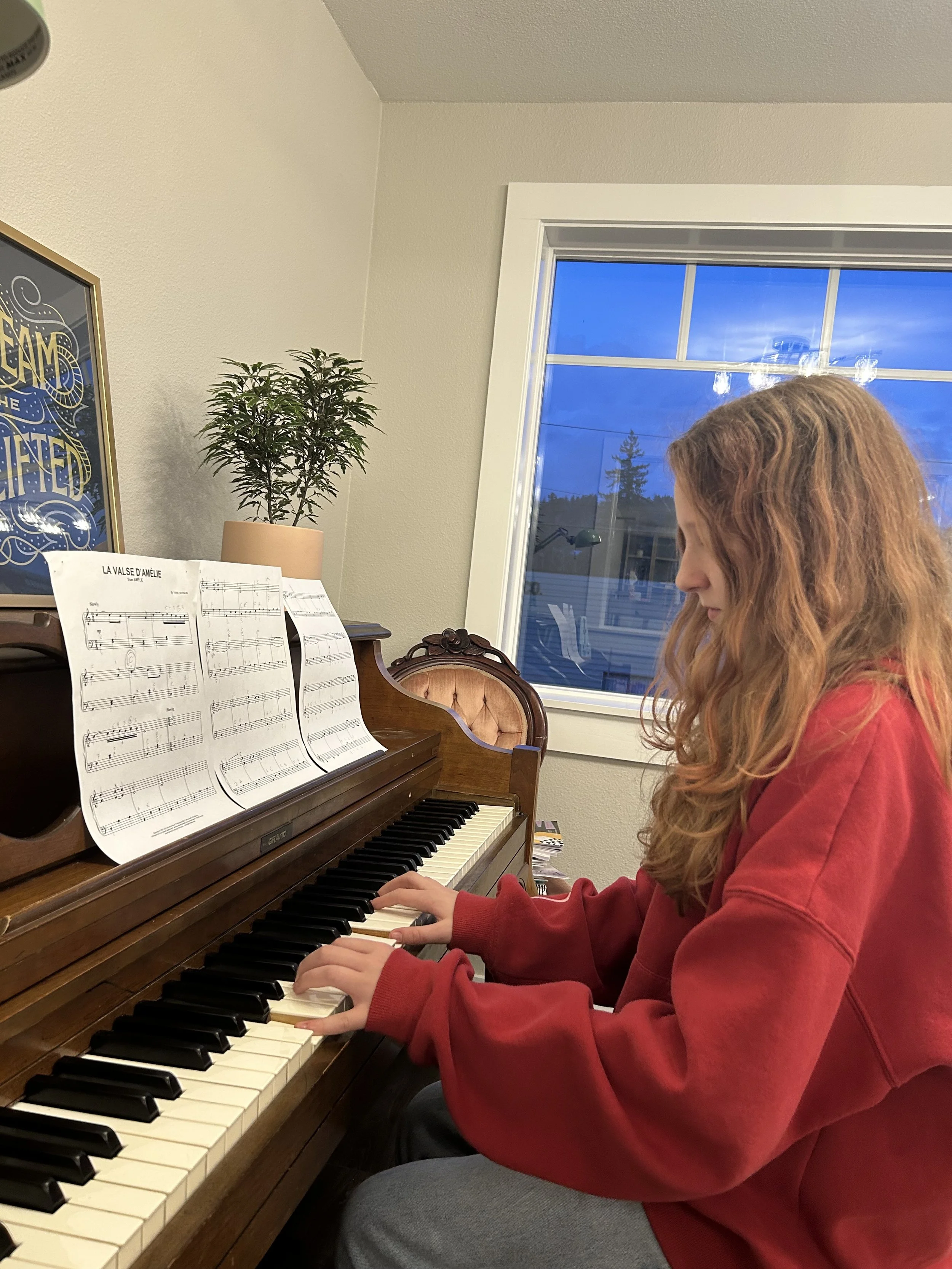 A young woman with curly red hair wearing a red hoodie playing a piano, with sheet music open in front of her, in a room with a window showing a blue sky and trees outside.