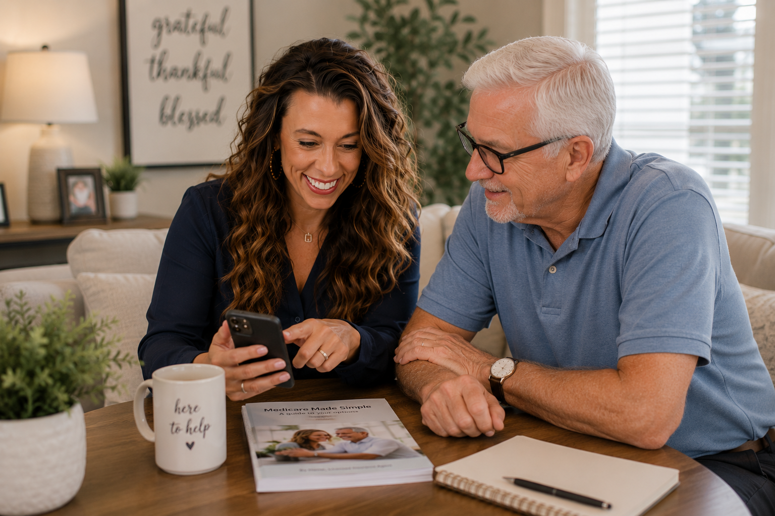 April and an older man sitting at a table, looking at a smartphone together and smiling, with paperwork and a mug on the table, in a cozy living room with a lamp, picture frames, and a large window.