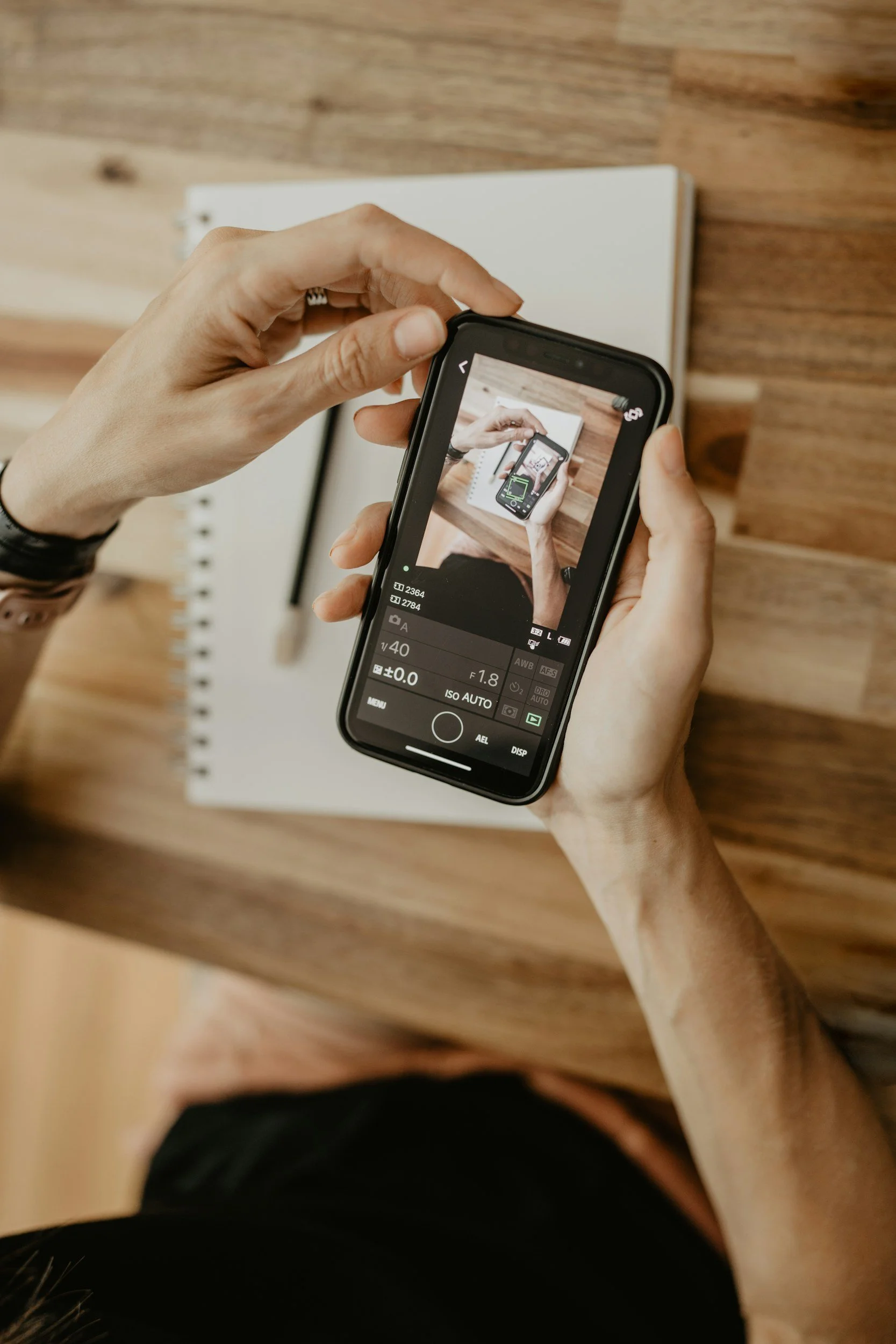 A person taking a photo of their phone screen with a camera app, showing hands holding the phone, in front of a wooden surface with a notebook behind it.