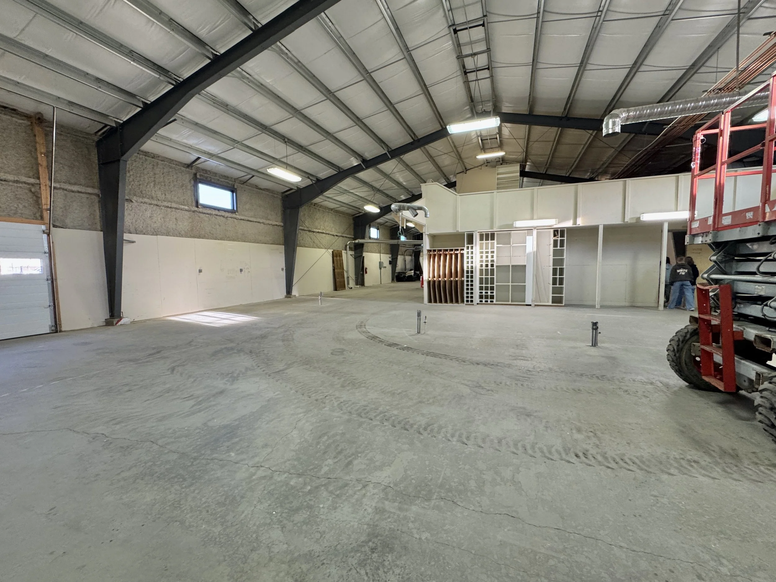 Interior of a large warehouse with exposed metal beams, concrete floors, some construction materials, and workers in the background.