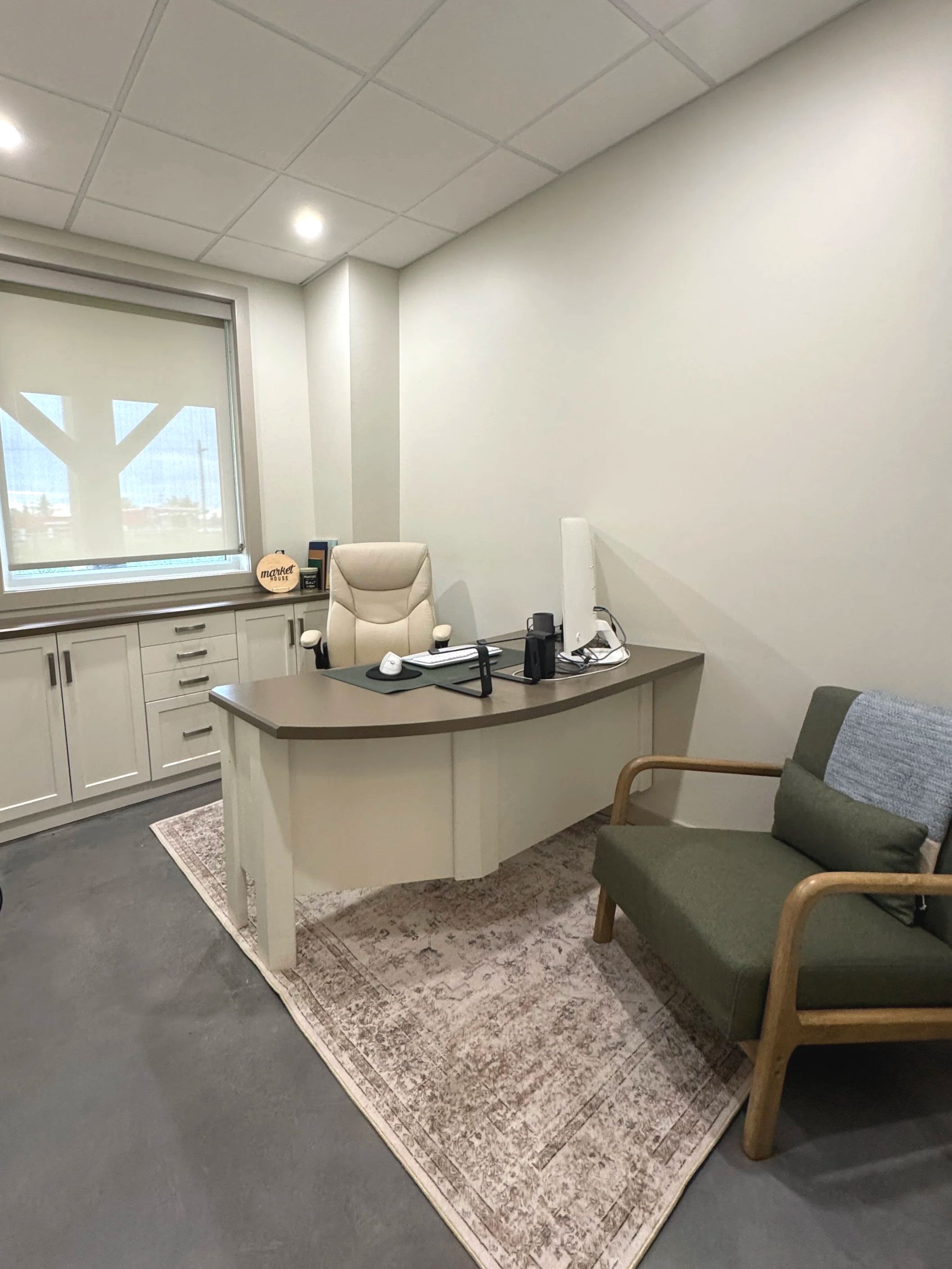 An office space with a desk, a white leather chair, and a green armchair. There is a window with a tree pattern on the blinds, white cabinets with drawers, and a patterned rug on the grey floor. Items on the desk include a computer monitor, mouse, and keyboard.