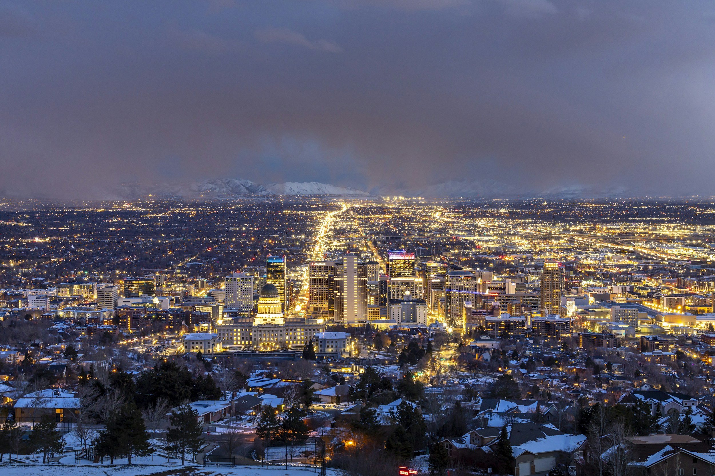 Nighttime cityscape with illuminated buildings, snow-covered rooftops, and distant snow-capped mountains under a cloudy sky.