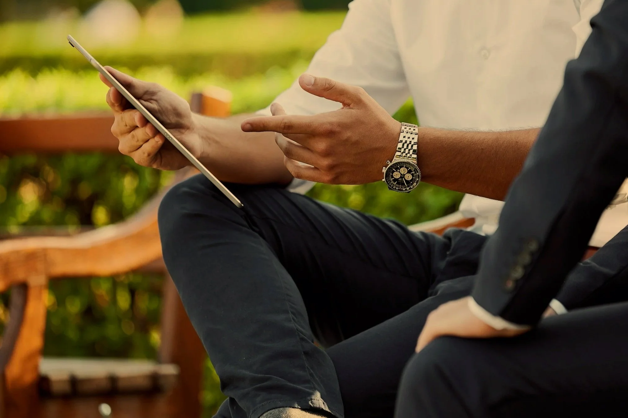 Close-up of two people sitting outdoors, one showing a smartphone and the other pointing at the screen. One person wears a silver wristwatch. The scene shows blurred greenery and a wooden bench in the background.