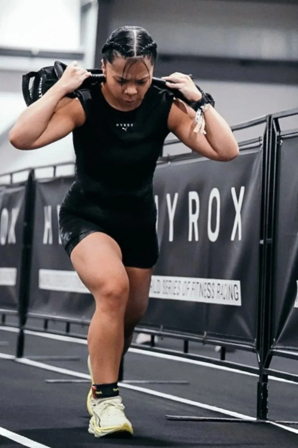 A woman wearing a black athletic outfit and yellow running shoes is carrying a sandbag on her shoulders during a workout or fitness competition in an indoor gym. She appears focused and determined.