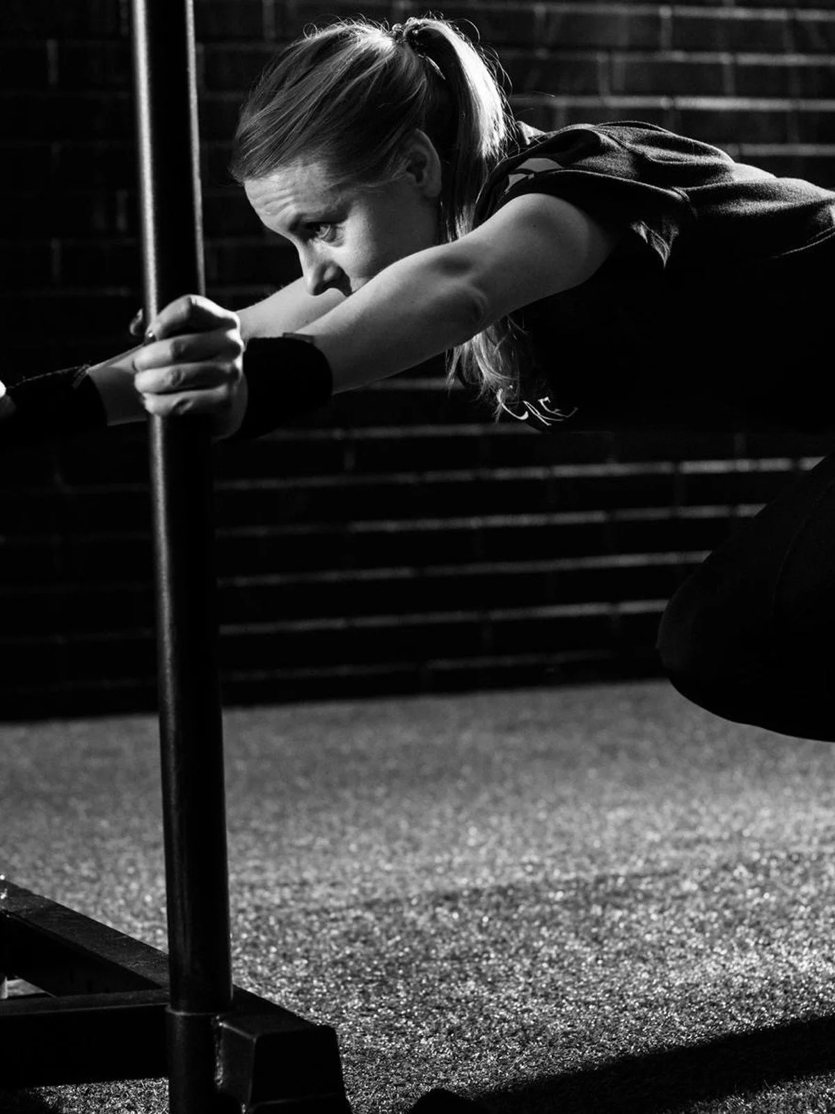 A woman with long hair in a ponytail performing a plank exercise with her arms extended and holding a pole, in a gym with a dark background.