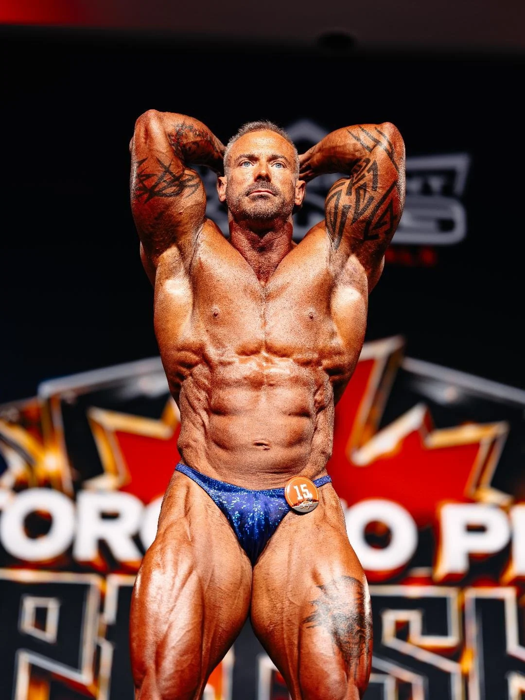 Male bodybuilder on stage posing with hands behind head, showing muscular physique and tattoos, wearing blue posing trunks with number 15, at a bodybuilding competition.