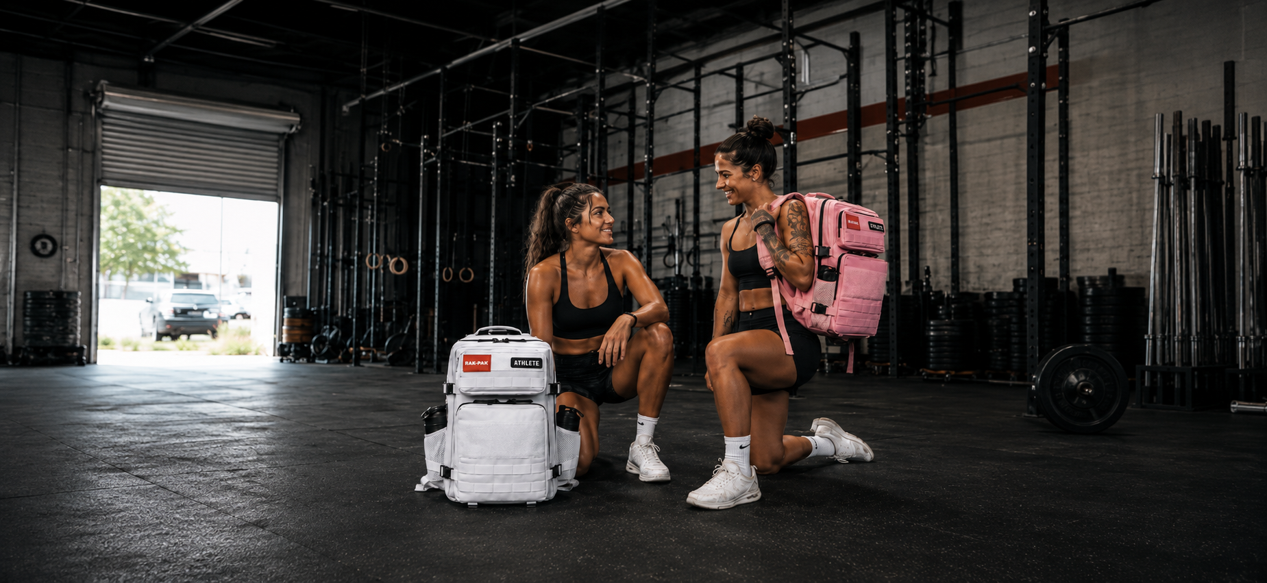 Two women in athletic black outfits inside a gym, kneeling and talking. One is holding a pink backpack, and two white backpacks with labels are on the ground. The gym has black rubber flooring, gym equipment, and an open garage door showing outside greenery and parked cars.