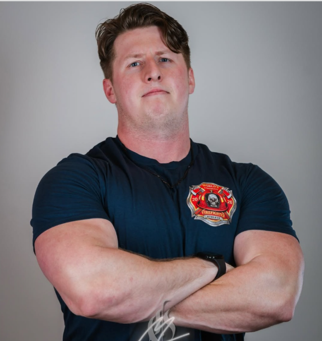A young man with crossed arms, wearing a firefighter t-shirt with a firefighter badge patch, looking confidently at the camera against a plain gray background.