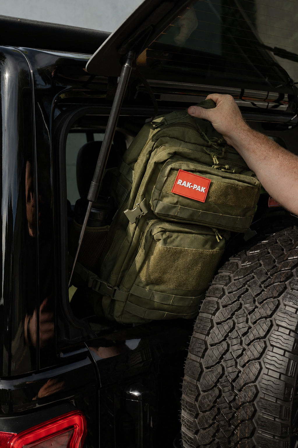 A person loading a green military-style backpack with a red RAK-PAK patch into the back of a black off-road vehicle with a spare tire mounted on the side.