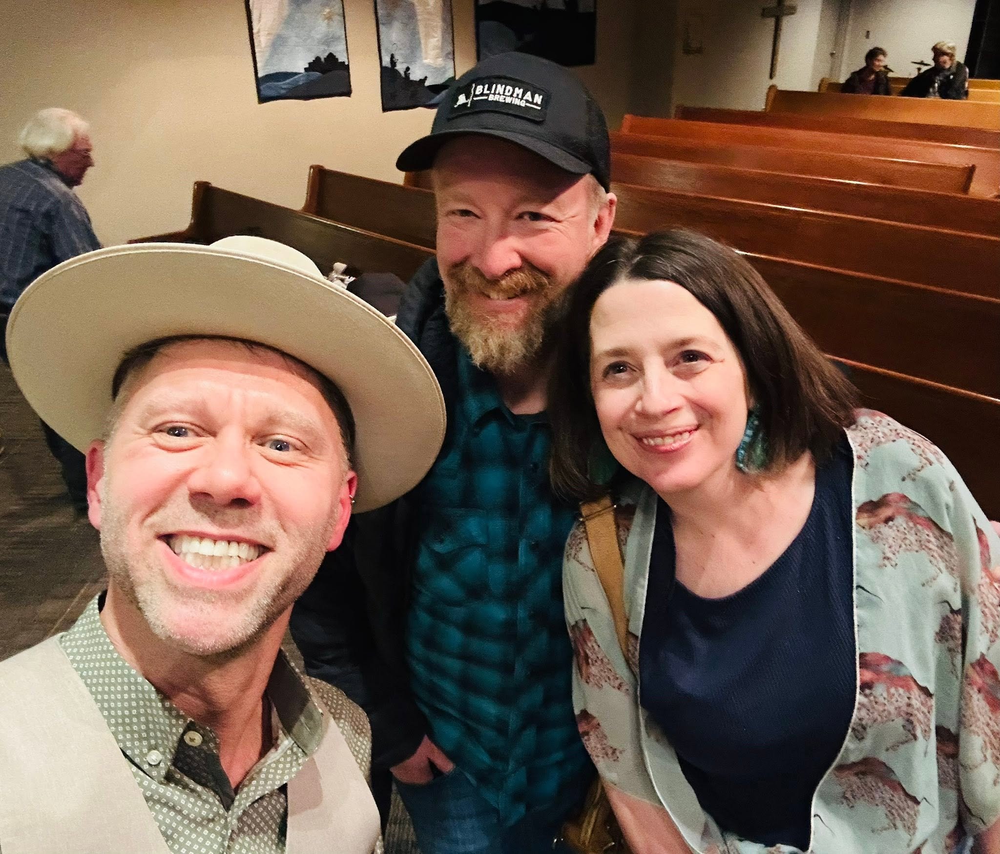 Group of four smiling people posing closely together indoors, with three individuals visible and two in the background, sitting on wooden pews, with artwork on the wall behind them.