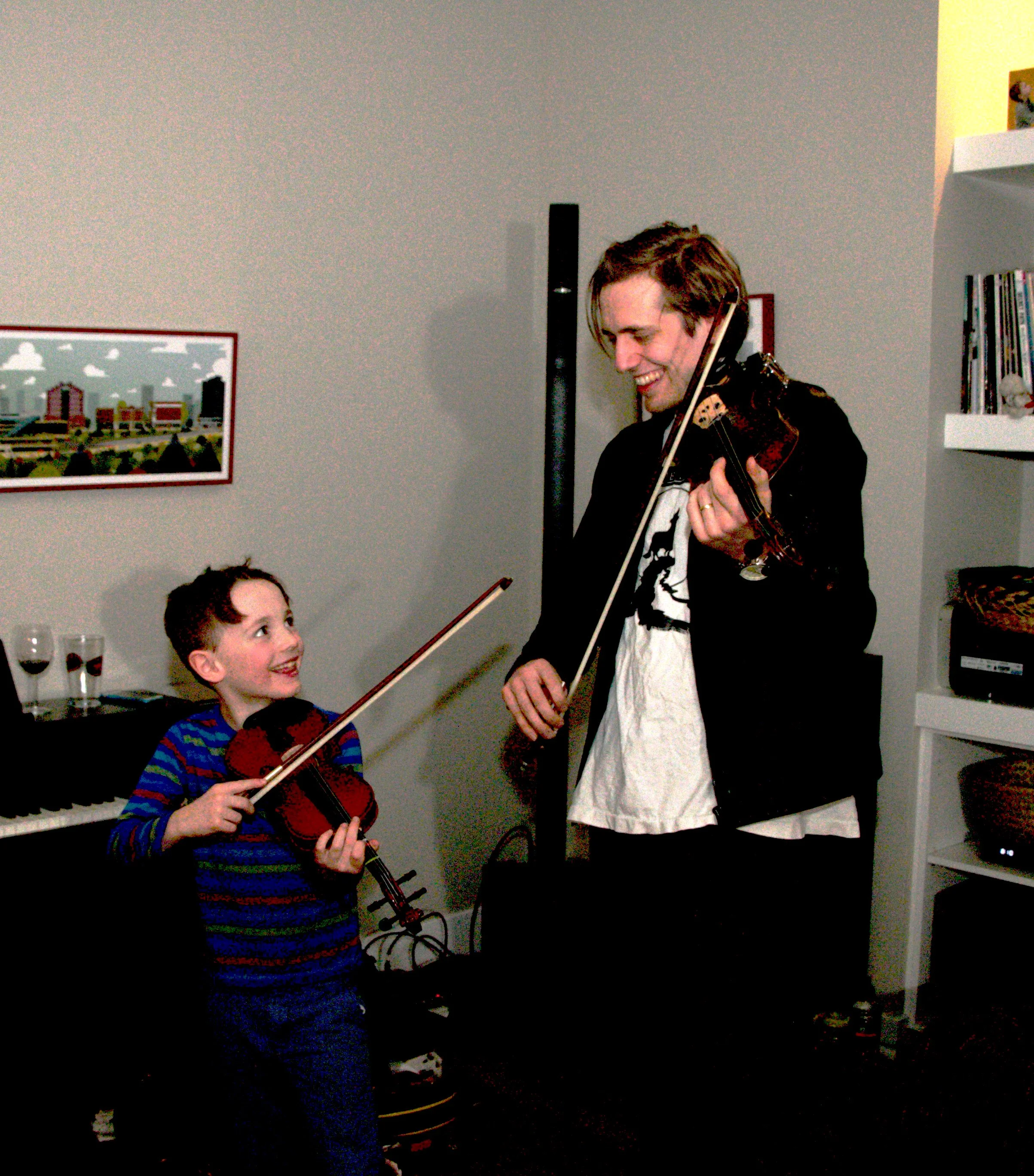 A young boy and an adult playing violins together indoors, smiling at each other, with books and a picture on the wall in the background.