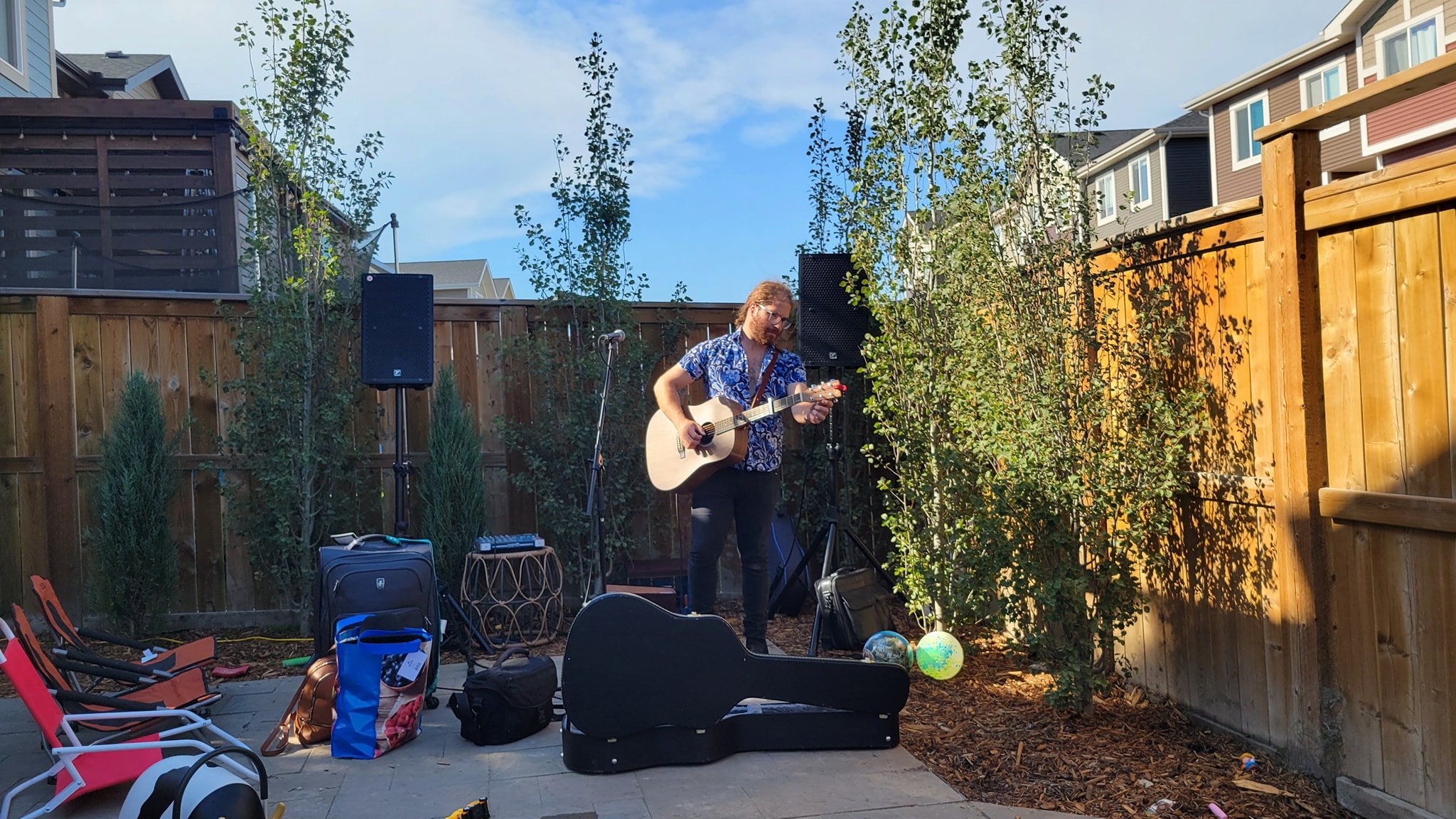 A man with glasses and a floral shirt playing an acoustic guitar at an outdoor backyard concert with speakers and musical equipment, surrounded by trees and houses.