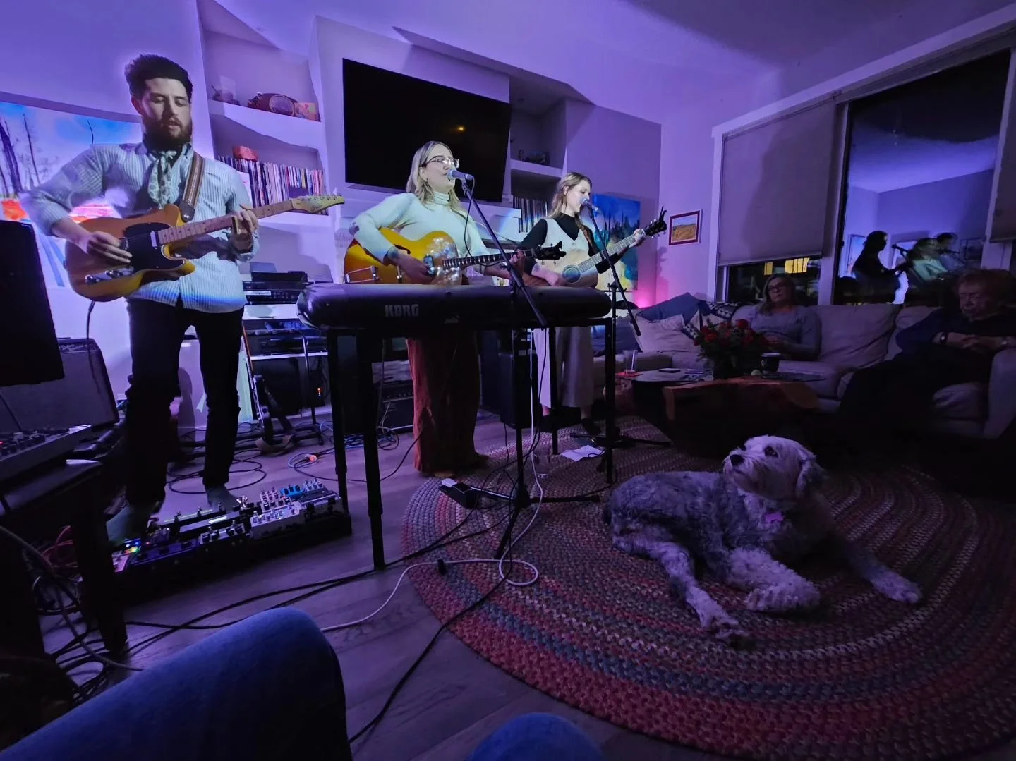 A group of four musicians performing in a living room with purple lighting. Three women and one man are playing guitars and a keyboard, singing into microphones. There are two women sitting on a couch watching, along with a man with gray hair and a w