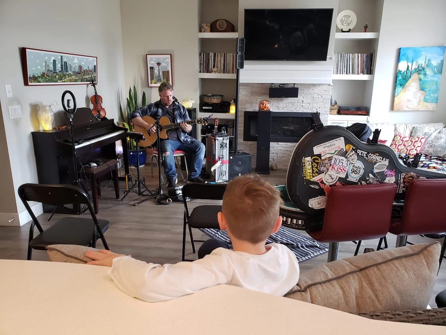 A man playing acoustic guitar in a living room with a young boy watching, several chairs, a covered guitar case with stickers, a piano, artwork, and a fireplace.