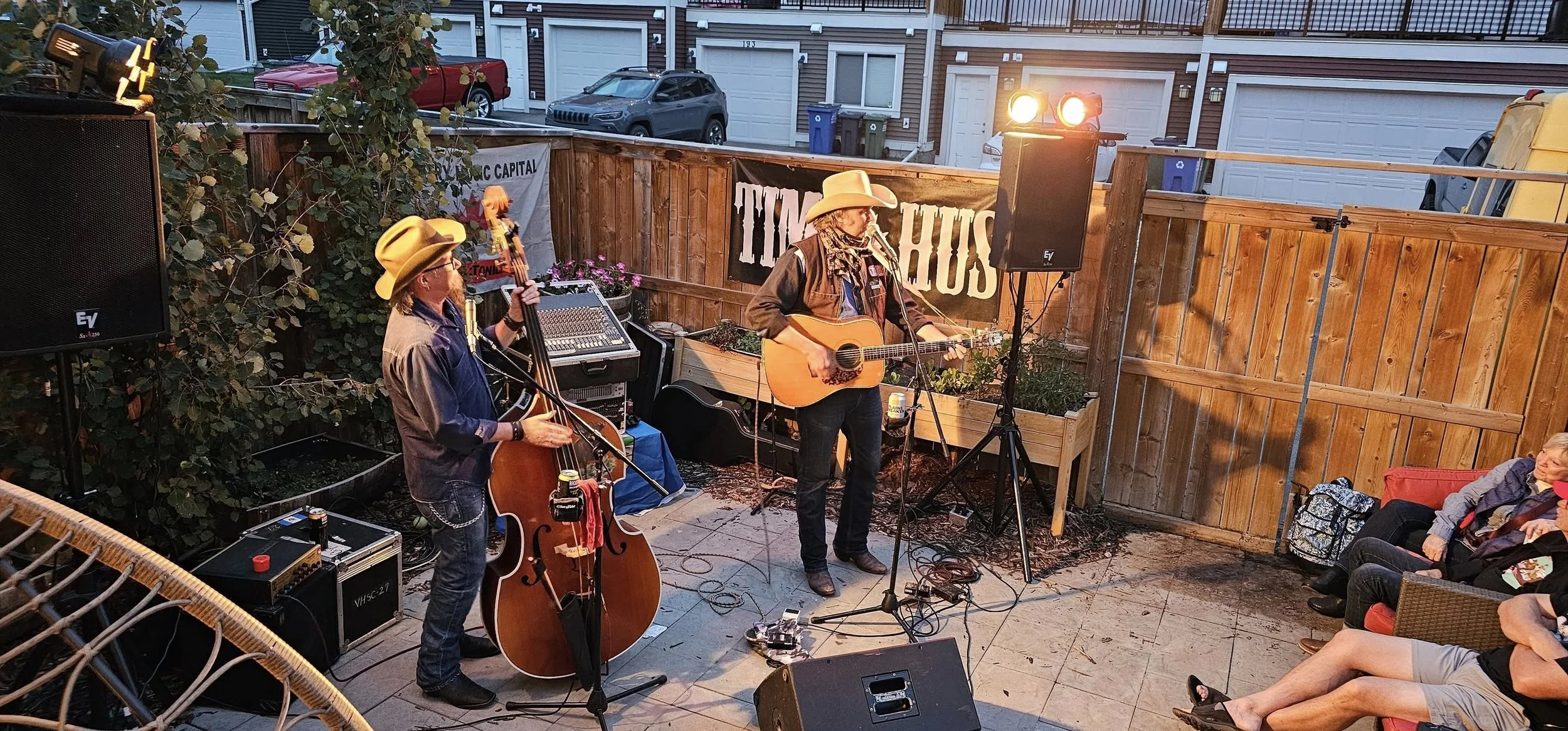 Two musicians perform with guitars, one also plays a double bass, on an outdoor patio with a small audience seated. The setting appears to be in a backyard, with a wooden fence and residential buildings in the background. The scene is lit with stage 