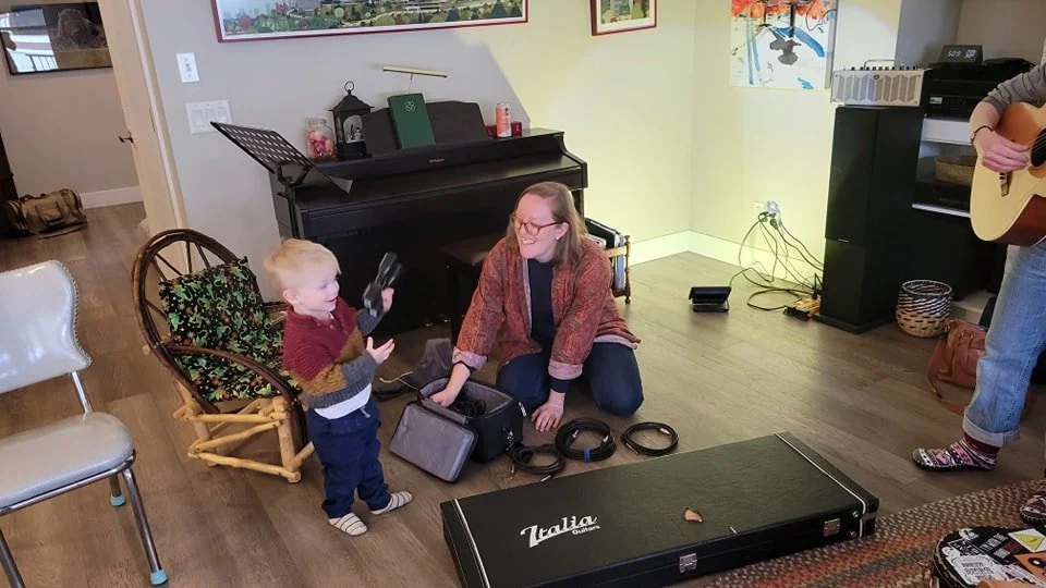 A woman and a young boy are inside a living room. The boy is interacting with a woman who is sitting on the floor, smiling. The woman is surrounded by various musical equipment, including headphones and a guitar. The room features a piano, artwork, a